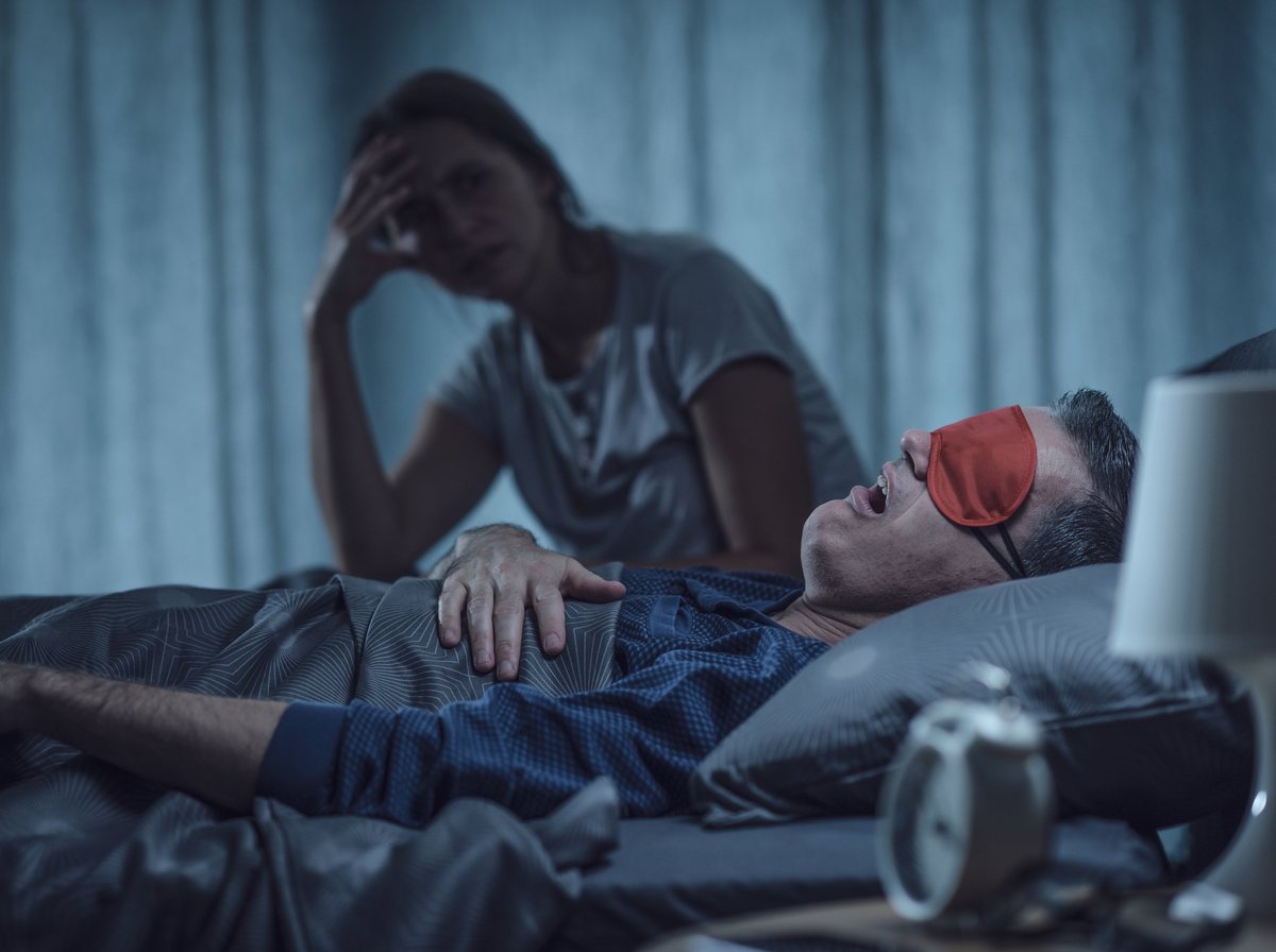 A middle-aged partner lies awake in bed at night with a subtle tired expression while their partner sleeps nearby