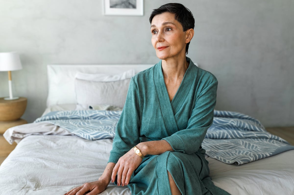 A woman in her fifties sits on the edge of a beautifully made bed in the early morning, gently touching the side of her neck