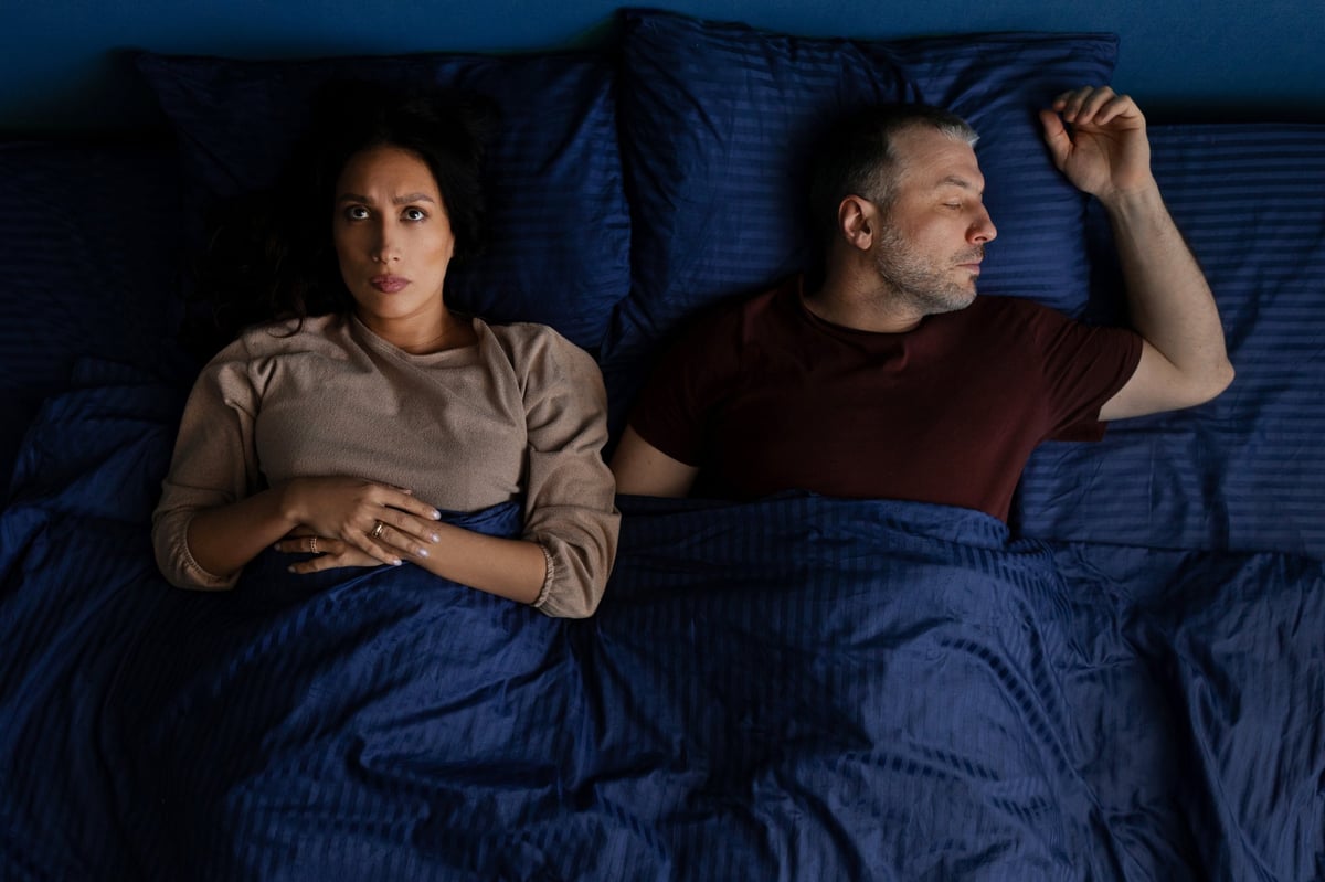 A top-down view of a middle-aged couple in bed at night: one partner is asleep while the other lies awake with a subtle tired expression