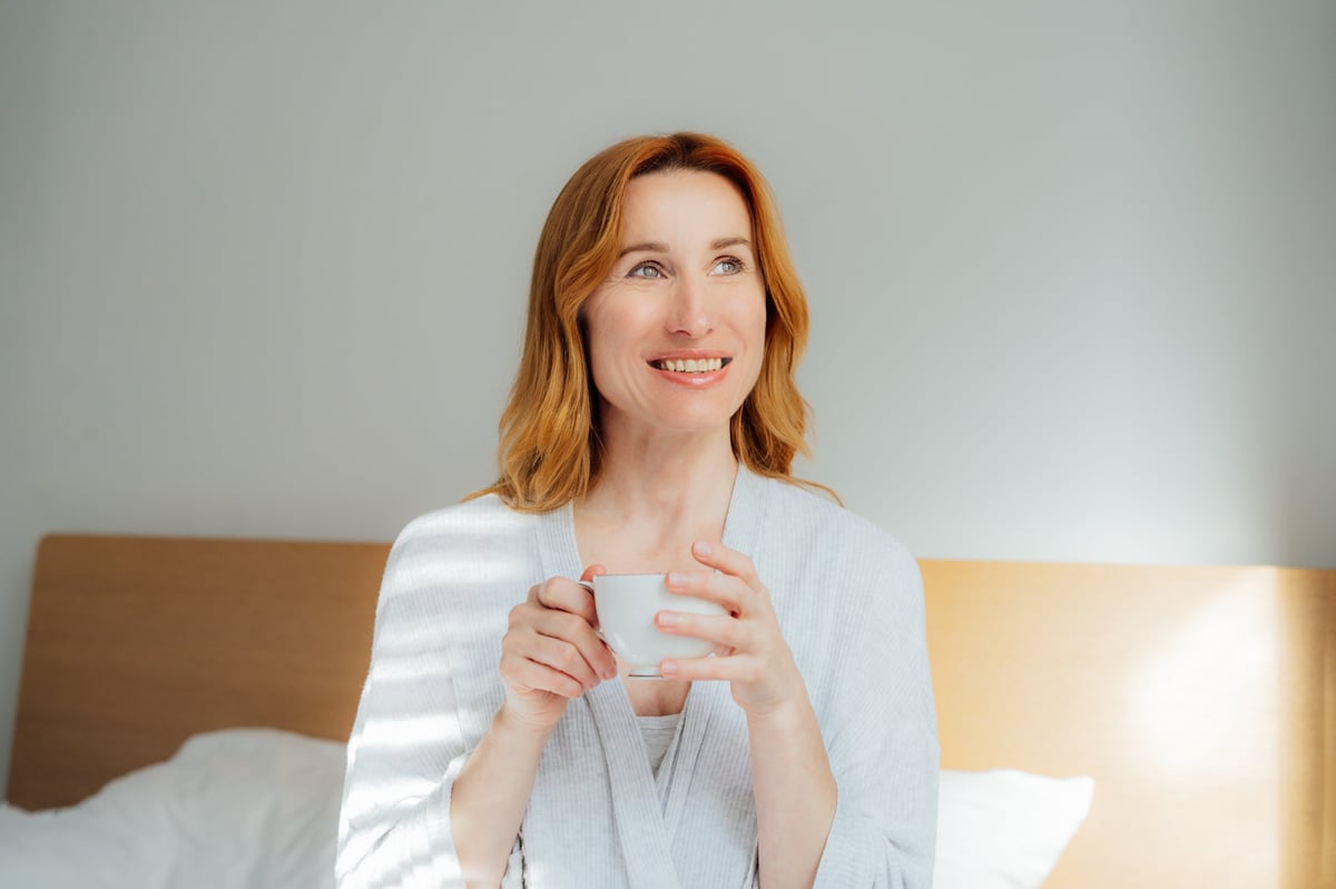 A woman in her fifties waking naturally in a bright modern bedroom, calm and comfortable